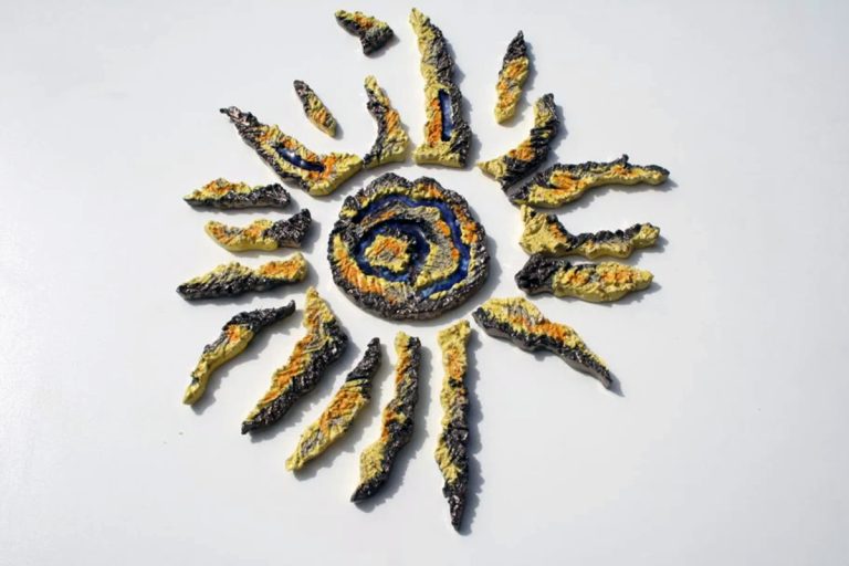 Scultura murale in ceramica bianca con fulcro centrale e raggi separati, decorata con graffi materici, tessere di vetro fuso e smalti giallo, arancio e oro