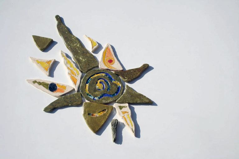Scultura ceramica di medio formato in argilla bianca con fulcro iridescente, vetri fusi e raggi radiali colorati in giallo, azzurro e argento