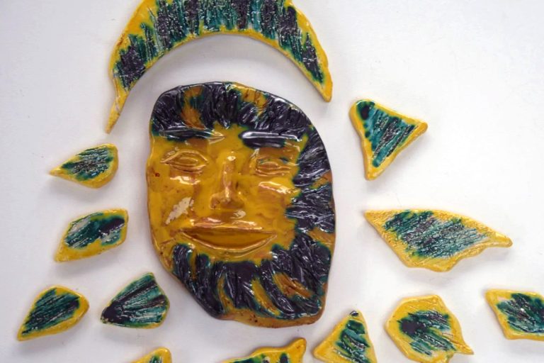 Sole in refrattario bianco con volto sorridente, raggi graffiati e finiture cangianti in smalto argento e giallo.