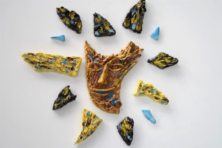 Scultura di sole in ceramica con maschera centrale in argilla rossa e un'alternanza di raggi materici neri, bianchi e azzurri.