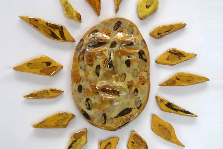 Sole in ceramica con maschera relazionale centrale, smalti gialli caldi e dettagli lucenti in foglia d'oro.