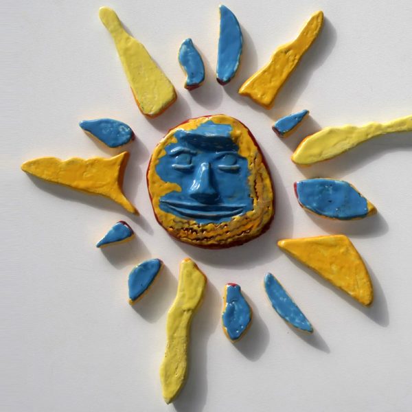 Sole in ceramica con volto astratto scolpito in arancione e azzurro, circondato da raggi gialli e azzurri.
