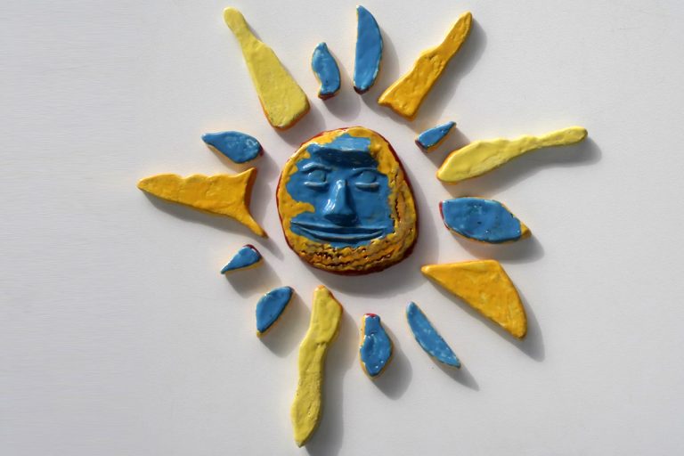 Sole in ceramica con volto astratto scolpito in arancione e azzurro, circondato da raggi gialli e azzurri.
