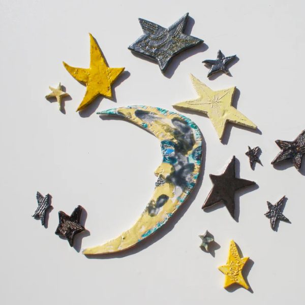 Piccola luna crescente in ceramica con un volto sorridente e spensierato, contornata da una costellazione di stelle.