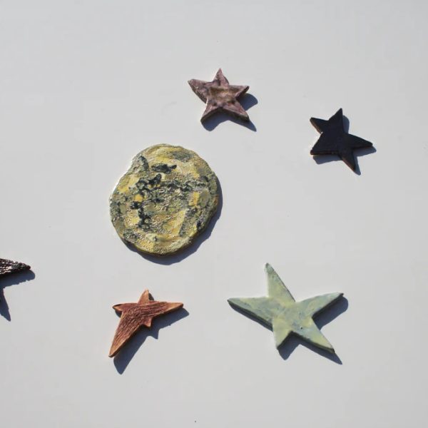 Luna scultorea piccola in ceramica bianca materica ricca di crateri, circondata da stelle artigianali colorate.