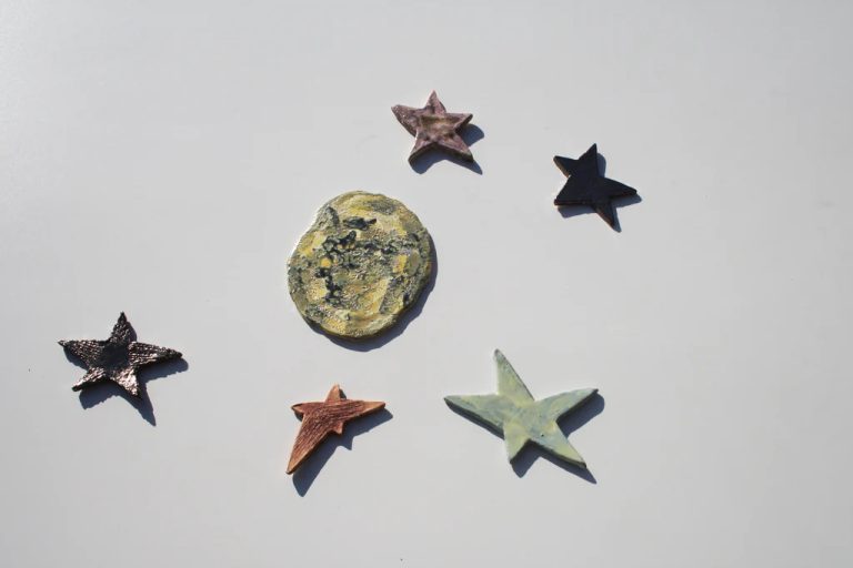 Luna scultorea piccola in ceramica bianca materica ricca di crateri, circondata da stelle artigianali colorate.