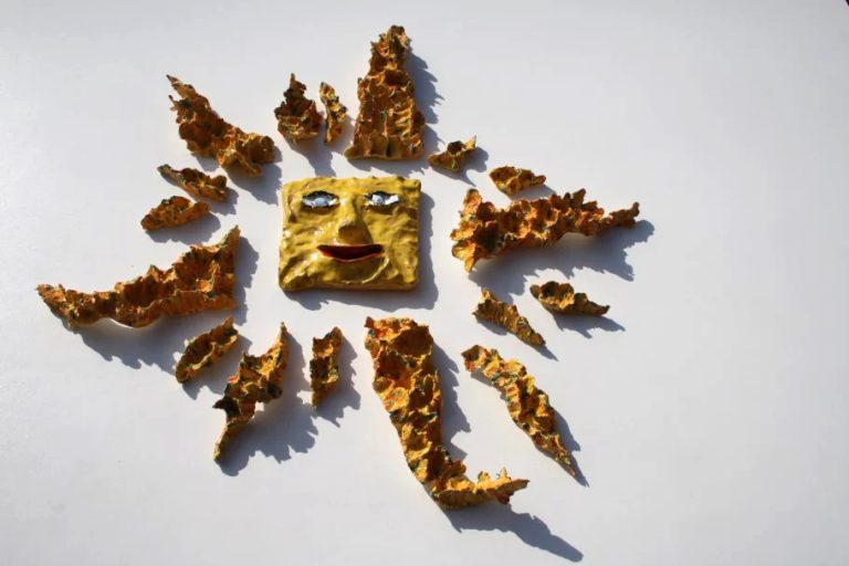 Sole medio-grande a rosa dei venti con volto sorridente centrale, raggi graffiati gialli, arancioni e verdi, e sette punte con inserti in vetro fuso.