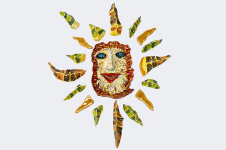 Piccolo sole a forma di Rosa dei Venti con maschera centrale sorridente gialla, fiamme rosse e diciassette raggi geometrici marroni, verdi e ad effetto.