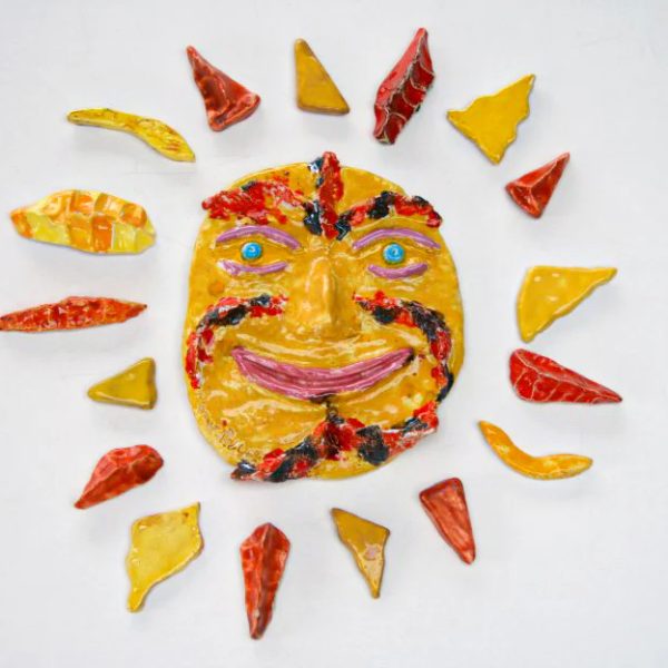 Piccolo sole giocherellone in argilla bianca con una maschera centrale sorridente e una ricca palette di smalti gialli e arancioni.